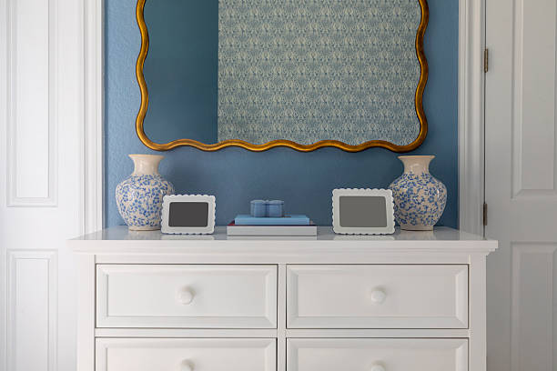 Elegant interior scene featuring a white dresser styled with blue and white ceramic vases, decorative photo frames, and stacked books, set against a soft blue wall. An ornate, gold-framed wall mirror adds a classic touch while reflecting subtle patterned textures.
