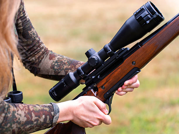 Hunter in camo gear looks closely at her rifle, adjusting or preparing it in rural setting. Mindful preparation, Woman and hunting. Gun safety and handling. Women in traditionally male roles
