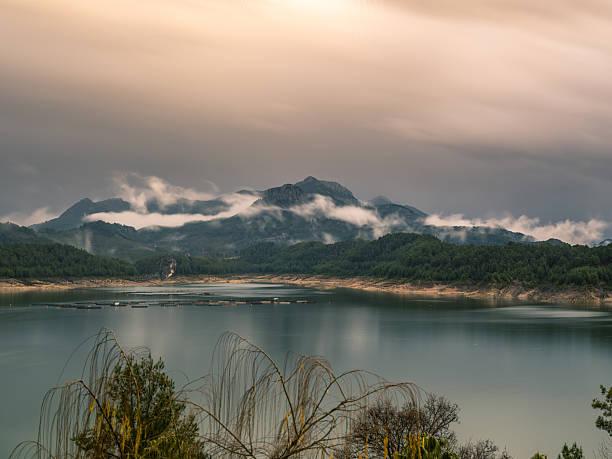 Wide angle view of a turquoise reservoir lake containing floating aquaculture fish cages. Pine tree covered mountains rise in the background with low lying mist and clouds clinging to the peaks under an overcast sky.