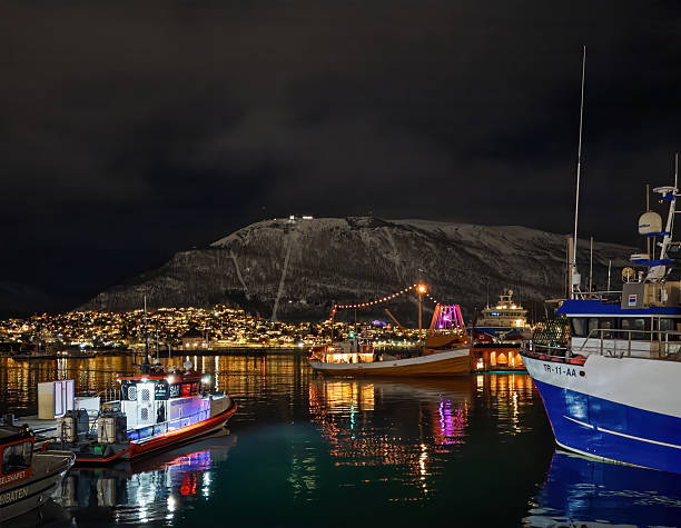 Tromso, Norway - December 21, 2025: Winter polar night. Looking east to the mainland with boats docked downtown. Black sky and many lights on houses and the boats, fishing and ferry