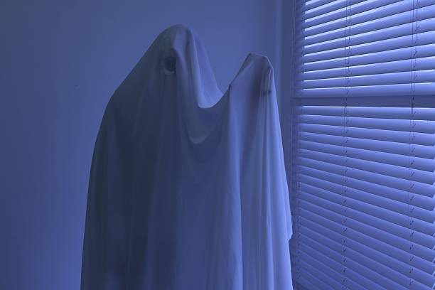 Sad ghost in white sheet outside the window of an abandoned house. Black and white photo