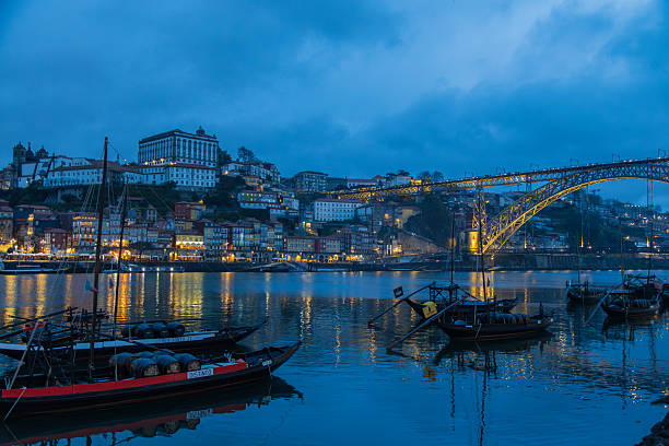 Porto, Portugal, 11 Jan 2026: Dom Luis I Bridge Over Douro With Illuminated Arch And Golden Lights, Waterfront Buildings Glowing, Traditional Boats Moored In Foreground, Historic Ribeira District