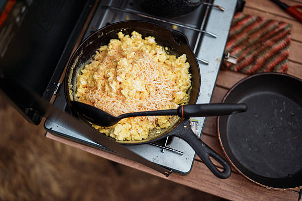 Scrambled eggs topped with shredded cheese cook in a cast iron skillet on a portable camp stove while breakfast is prepares at a riverside campsite.