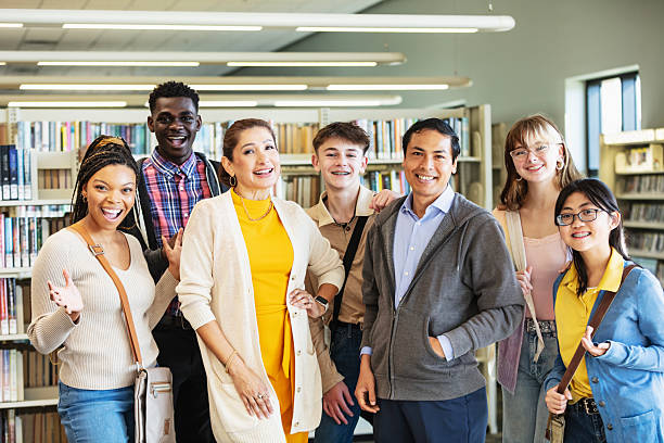 A multiracial group of five high school students and two teachers standing in the library together, smiling at the camera.