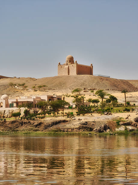 A breathtaking view of a serene river landscape with a historical monument atop a hill. Smaller houses and lush vegetation adorn the riverbanks, giving insight into the picturesque Egyptian scenery.