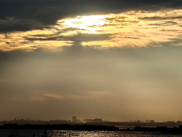 A breathtaking view of the sky at sunset or sunrise, where golden light breaks through thick clouds, casting dramatic rays across the atmosphere. The warm glow illuminates the horizon, highlighting the silhouette of a distant city skyline against a hazy backdrop. Below, calm water reflects the soft light, adding depth and tranquility to the scene. This atmospheric landscape captures the quiet beauty of nature meeting urban life, evoking feelings of peace, reflection, and awe. Ideal for themes such as natural light, cityscapes, twilight moments, or environmental harmony.