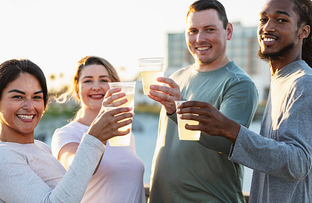 A multiracial group of four friends, in their 20s and 30s, standing together on a boardwalk at the beach, having refreshments. They are raising their cups of lemonade to toast their friendship, smiling at the camera.