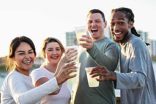 A multiracial group of four friends, in their 20s and 30s, standing together on a boardwalk at the beach, having refreshments. They are raising their cups of lemonade to toast their friendship, smiling, laughing and looking toward the camera.