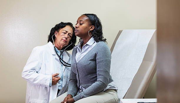 A mid adult African-American woman sitting on an exam table in a doctor's office, as the female doctor listens to her lungs with a stethoscope. The doctor is a multiracial senior woman wearing a white lab coat.