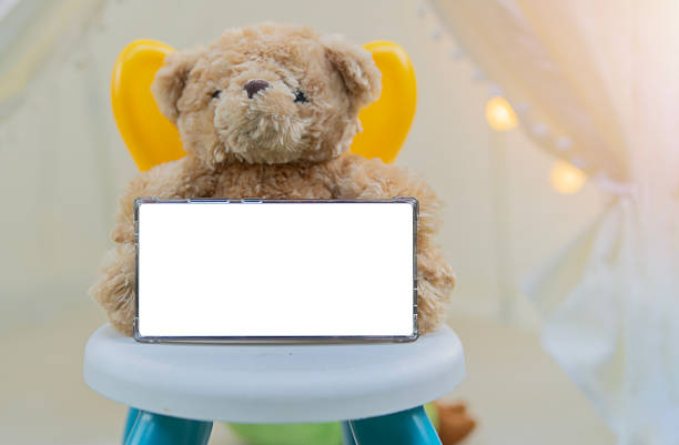 Teddy bear with phone mock up with isolate screen sitting on kid's chair over teepee tent or kid's playroom. Copy space and selective focus.