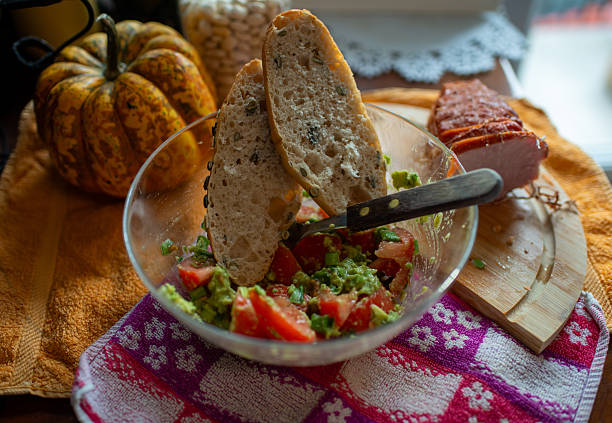 Glass bowl with mashed avocado and chopped tomatoes served with sliced rustic bread, pumpkin seeds, and smoked meat on a wooden board. The scene represents homemade food, breakfast, and casual home dining.