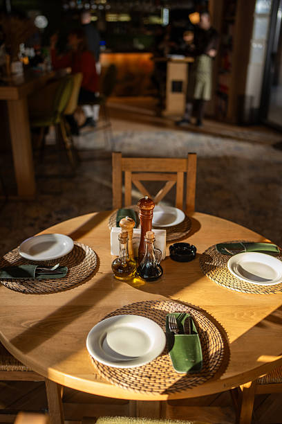 A cozy round wooden table sits ready for guests in a warmly lit restaurant. Plates, woven placemats, green napkins, and condiment bottles create an inviting, ready-to-dine atmosphere.
