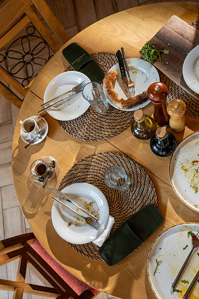 A round wooden table shows remnants from a meal, with empty plates, forks and knives, a crusty bread piece, condiments, and glassware, creating a warm, homey dining scene captured from above.