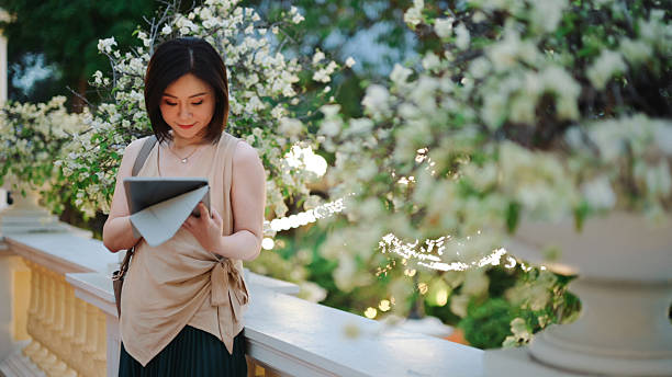 lifestyle footage expressing digital connection, mindfulness, and emotional balance asian woman standing outdoors while using tablet in peaceful environment. themes include personal time, technology use, calm lifestyle, and modern urban living.