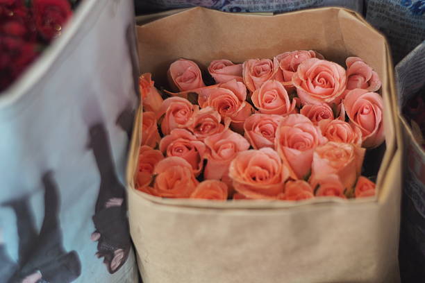 A vibrant bouquet of fresh, peach-pink roses bundled in simple brown kraft paper, ready for sale at a bustling flower market.