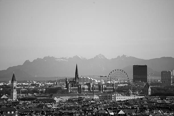 Black and white panoramic view of Munich city skyline featuring historic church towers, urban architecture, and the Bavarian Alps in the background. A timeless cityscape of Munich, Germany, captured from an elevated viewpoint with a blend of traditional and modern elements