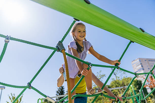 Young girl smiling, climbing on a green rope structure, her mother supporting her from below at an outdoor playground