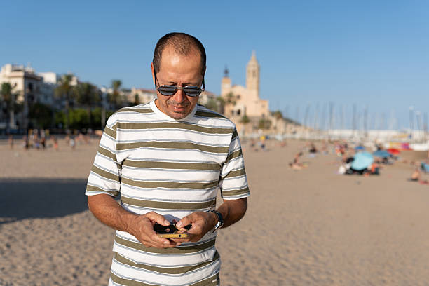 Middle-aged man using a mobile phone on a sandy beach under clear blue sky, enjoying travel