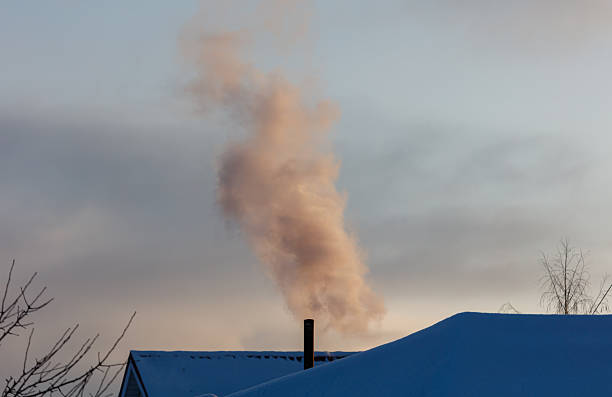 A chimney is smoking and the smoke is rising into the sky. The sky is cloudy and the sun is setting