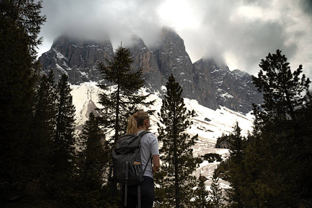 Woman with a backpack standing in a forest and looking toward steep rocky mountains with snow and clouds