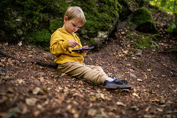 A cute blond toddler, around two to three years old, is sitting on the ground outdoors and looking at a mobile phone. Dressed in comfortable clothes suitable for outdoor activities, the child appears calm and curious while focusing on the screen. The scene captures early childhood, curiosity, modern family life, and the presence of technology in everyday moments, set in a natural environment.