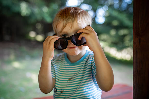 A portrait of an adorable blond toddler, around two to three years old, wearing a T-shirt stained with chocolate while playing outdoors. The child is smiling and putting on oversized adult sunglasses, which look too big for him and add a playful, humorous touch. The scene captures childhood innocence, curiosity, messy play, fun imagination, and joyful everyday moments in early childhood.