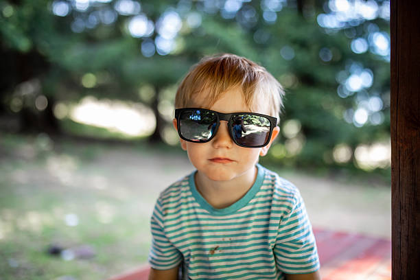 A portrait of an adorable blond toddler, around two to three years old, wearing a T-shirt stained with chocolate while playing outdoors. The child is smiling and putting on oversized adult sunglasses, which look too big for him and add a playful, humorous touch. The scene captures childhood innocence, curiosity, messy play, fun imagination, and joyful everyday moments in early childhood.