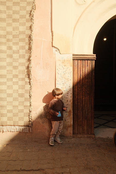 One young child standing against a wall and woven rug in golden hour light, reflecting childhood in Morocco