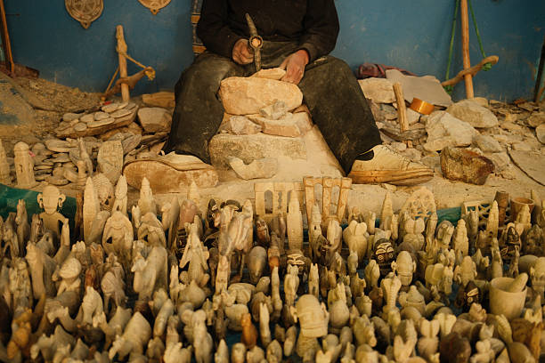 Craftsman using hammer and chisel, shaping stone into various small figurines. Displaying many finished handcrafted pieces