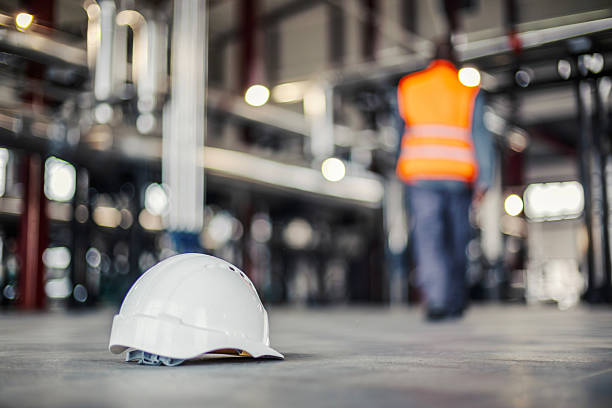 White hard hat resting on a concrete floor in a factory with an unrecognizable worker in an orange vest walking away in the blurred background, illustrating labor completion or a break