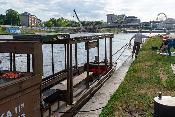 Krakow, Poland – June 29, 2025: A wooden ferry is moored at the riverbank while people prepare equipment along the shore, with construction cranes and urban buildings visible across the river.