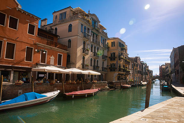 Sunlit view of a traditional canal in Venice, lined with historic residential buildings, balconies, and small boats moored along the waterway. The clear daylight highlights the warm colors of the facades and the calm, everyday character of this quiet Venetian neighborhood, offering an authentic glimpse of local life away from iconic landmarks.