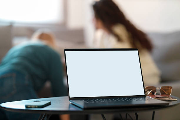 Laptop computer with a blank white screen on a table indoors at home, with two women blurred in the background.
