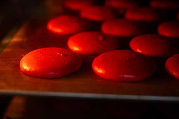 Red macarons baking on a tray inside a hot oven, showing the confectionery slowly rising and developing their characteristic feet in the warmth, creating a delicious gourmet dessert