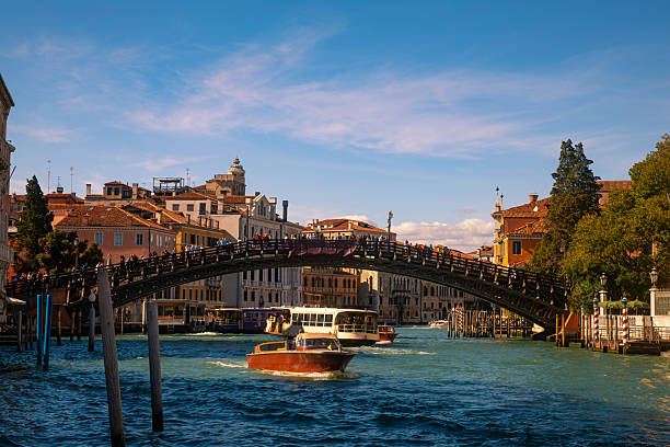 Lively view of a main canal in Venice, Italy, featuring a historic arched bridge crowded with pedestrians and boats navigating the waterway below. The image captures the everyday movement of the city, with traditional architecture, wooden mooring poles, and active water traffic reflecting the dynamic rhythm of Venice.