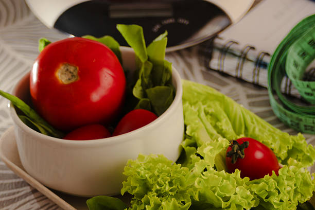 Fresh red tomatoes and crisp lettuce in a white ceramic bowl beside a kitchen scale, spiral notebook and green measuring tape, symbolizing meal planning, weight loss and healthy living