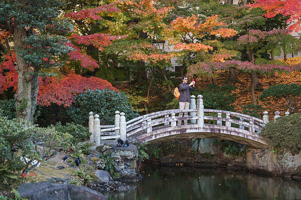 A man standing on a stone bridge in a public park during autumn, holding a smartphone and taking a photo. He is wearing casual clothing and a backpack, surrounded by colorful fall foliage, trees, and a calm pond below the bridge. The scene shows a single adult male using a mobile phone in a natural landscaped park environment during daytime, with seasonal atmosphere and copy space visible.