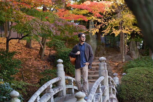 A man walking across a stone bridge in a public park during autumn. He is wearing smart casual clothing and carrying a backpack, surrounded by colorful fall foliage, trees, and landscaped greenery. The scene shows a single adult male in a natural urban park environment during daytime, with a pond below the bridge and seasonal atmosphere visible, offering copy space.