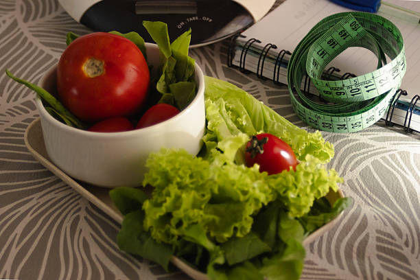Fresh vegetables, including lettuce and tomatoes, sit in bowls and on a plate with a measuring tape and notebook, symbolizing a commitment to healthy eating, diet planning, and active weight loss