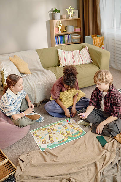 Group of three Generation Alpha kids, including Caucasian boy, Caucasian girl, Black girl, sitting on floor playing board game together, using digital gadget nearby