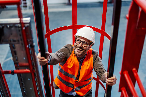 Smiling man wearing hard hat and safety vest climbing a caged industrial ladder inside a modern factory or construction site, representing success and progress