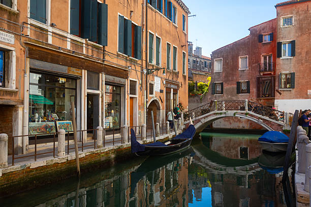 Sunlit view of a traditional canal in Venice, lined with historic residential buildings, balconies, and small boats moored along the waterway. The clear daylight highlights the warm colors of the facades and the calm, everyday character of this quiet Venetian neighborhood, offering an authentic glimpse of local life away from iconic landmarks.