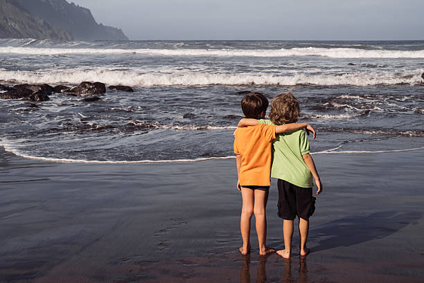 Two young boys, brothers or friends, stand hugging on beach with black volcanic sand, Atlantic Ocean. Unrecognizable children, friendship, family bond, childhood, traveling in Spain, Canary Island