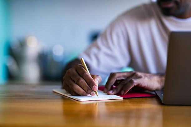 A person writes on a small notebook beside a laptop, capturing concentration and creativity intently.