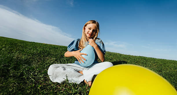 A young girl sits on a grassy field, holding a light blue balloon while a large yellow ball rests nearby. Clear blue sky and a relaxed, playful outdoor moment.