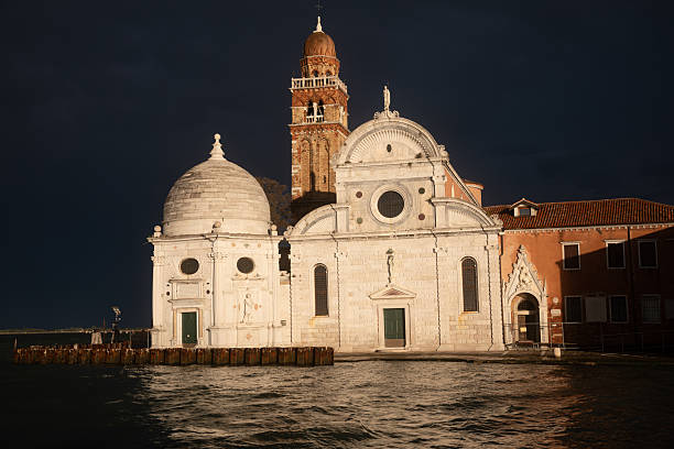Dramatic daylight view of the Church of San Michele in Isola, located on San Michele Cemetery Island in the Venetian Lagoon. After a stormy and overcast moment, sunlight breaks through the clouds and illuminates the historic Renaissance church, creating strong contrast between light and dark sky.