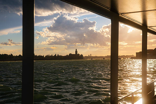 Dramatic sunset over the Venice Lagoon seen from a moving boat, with golden light reflecting on rough water and towering clouds illuminated after a storm. The historic skyline of Venice appears in silhouette under changing weather conditions, creating a powerful atmosphere that combines travel, nature, and urban landscape.
