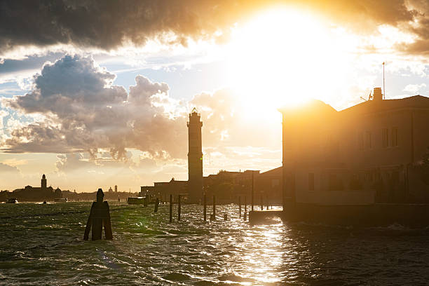 Golden sunset light breaks through dramatic storm clouds over the Venice Lagoon, illuminating the Murano Lighthouse and surrounding buildings. Rough water reflects the intense sunlight, creating a powerful atmosphere that highlights Venice’s relationship with weather, water, and navigation.