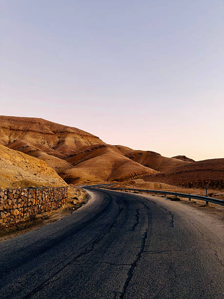 Jordan Ma’in Valley Mountain Winding cracked asphalt road curving through arid desert Ma’in Mountains and Hills under a soft evening sky, representing travel, adventure, and exploration. Dead Sea, Jordan, Middle East