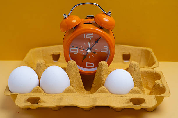 Orange alarm clock with fresh white eggs in yellow carton box. Morning routine and breakfast timing concept.
